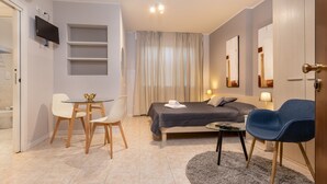 Une chambre d’hôtel moderne, équipée d’un lit, d’un bureau avec des chaises, d’une télévision et d’une salle de bain visible à travers une porte ouverte.