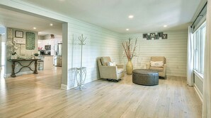 A spacious living room with a wooden floor, a sofa, a chair, and a coffee table. There is a kitchen in the background with white cabinets.