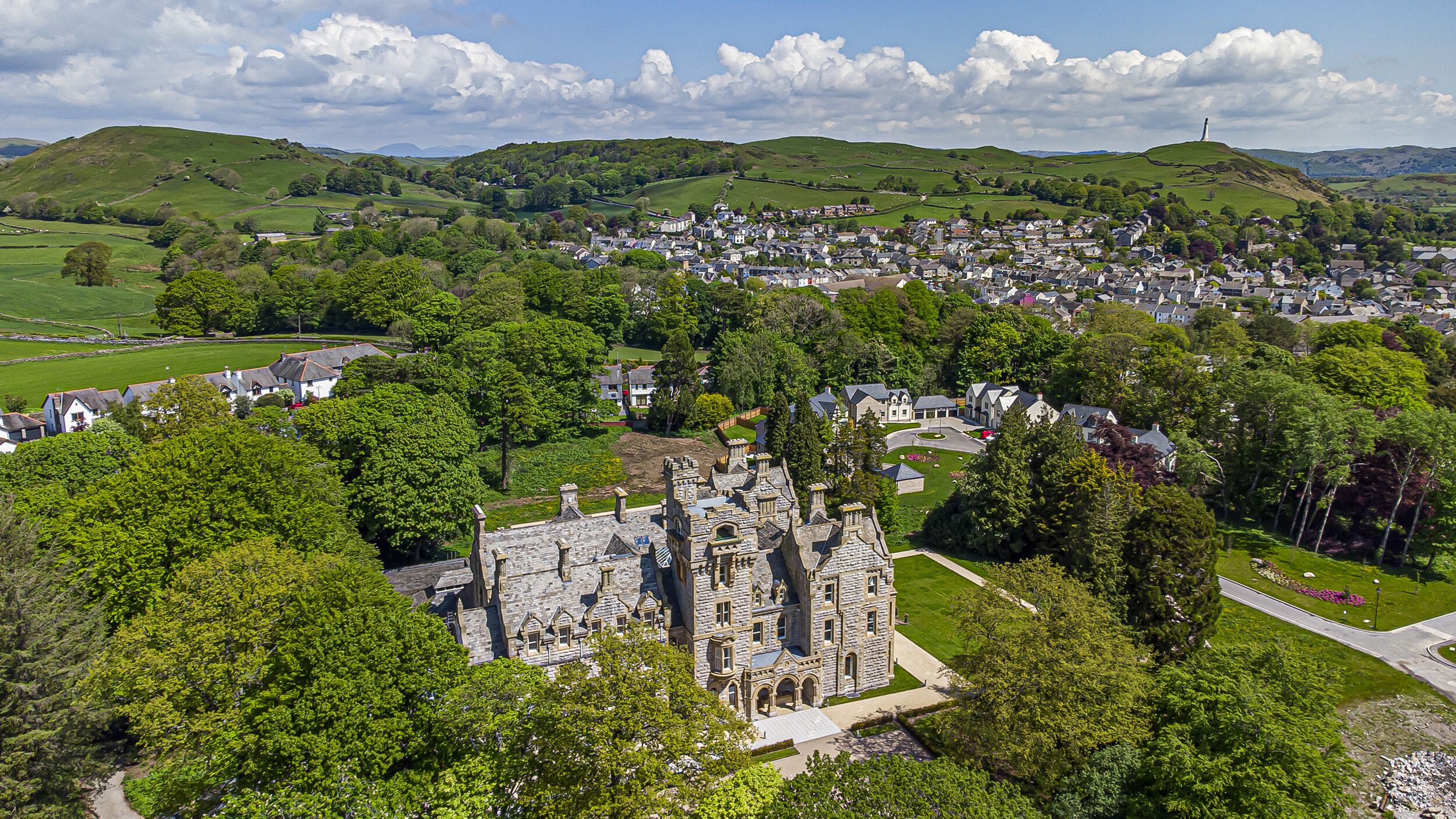 A historic stone building with a prominent tower, surrounded by lush greenery and a small village in the background.
