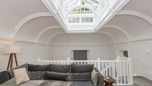 A spacious room with a high ceiling and a skylight, a grey sofa with cushions, a small wooden side table, and a white railing. - The Kennedy House Stone Cross Mansion (Ulverston)
