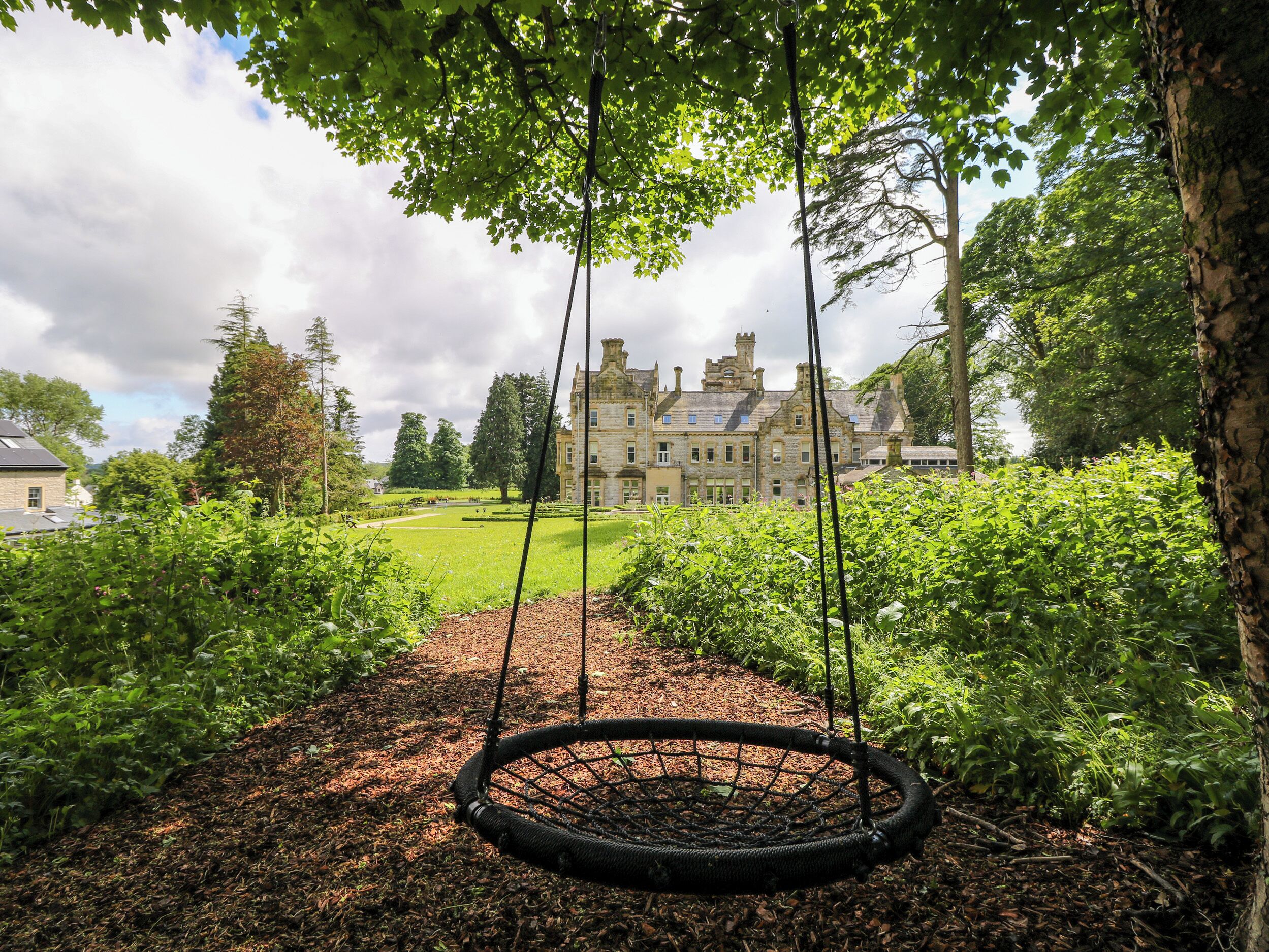 A garden with a swing set, a lawn, and a large building in the background.