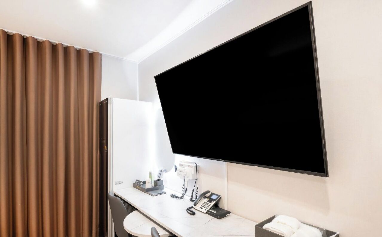 A hotel room with a flat-screen TV mounted on the wall, a desk with a telephone, and a minibar.