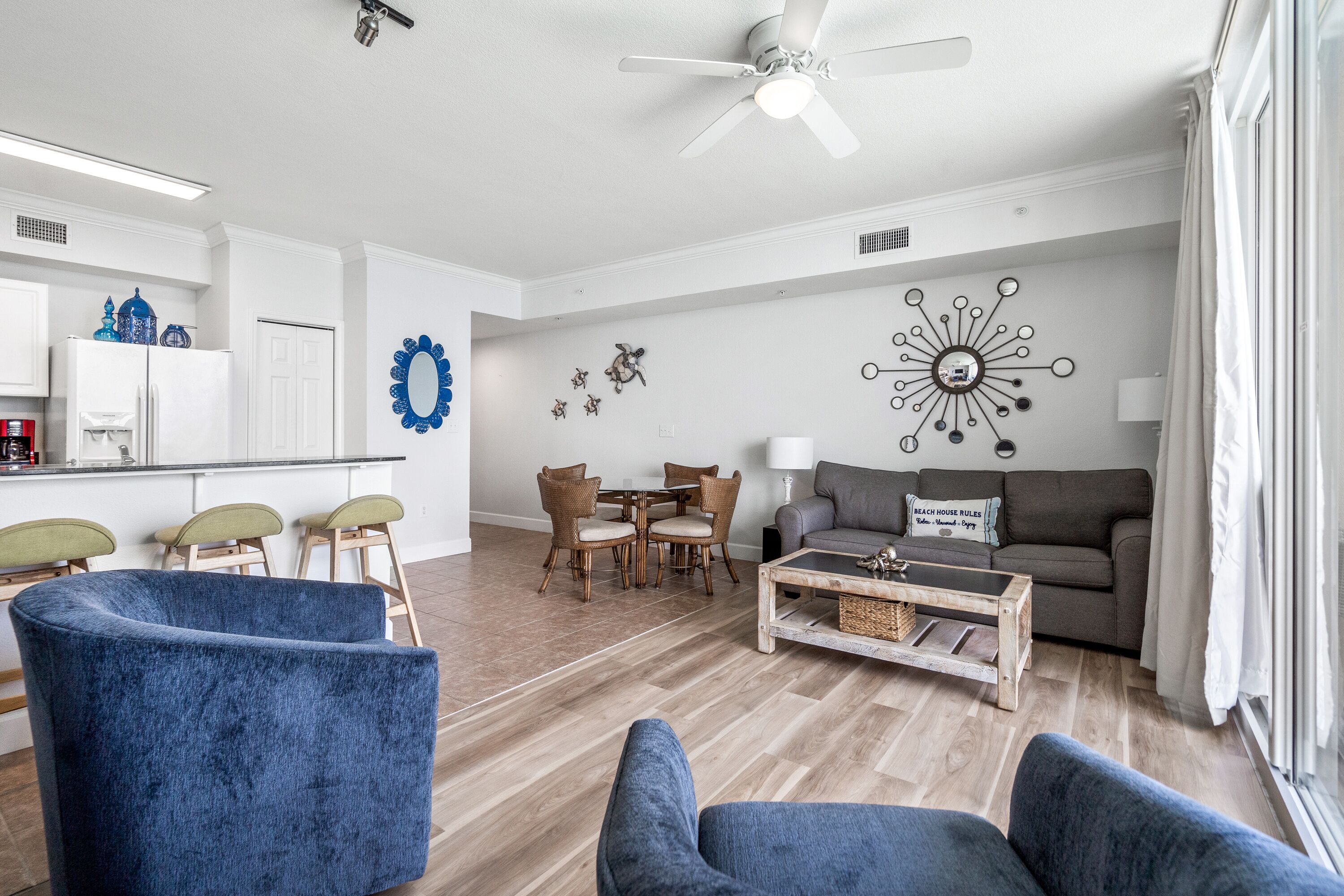 Un salon moderne doté d’un ventilateur de plafond, un coin repas avec des chaises en osier et une cuisine avec des meubles blancs.