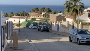 Rua com carros estacionados, uma palmeira e edifícios ao fundo, com vista para o mar.