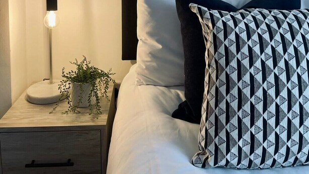 Un lit bien fait, avec une tête de lit noire, des oreillers blancs à motifs, une table de chevet avec une lampe et une petite plante en pot.