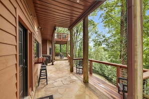 A wooden cabin with a stone patio, outdoor seating, and a covered area overlooking a forest.