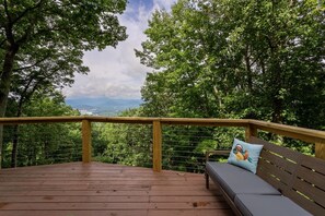 Um deck de madeira com um banco e almofada, cercado por árvores e vista para montanhas distantes.