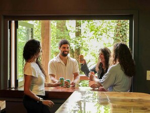 Un grupo de personas alrededor de una mesa de madera con bebidas, en una habitación con un ventanal grande que da a un bosque.