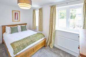 A bedroom with a wooden bed, green pillows, a white duvet, a green throw, a window with curtains, a radiator, and a picture on the wall.