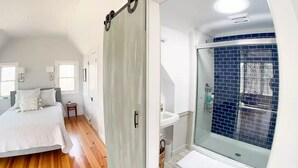 Une chambre avec un plancher en bois, un lit, une table de chevet et une porte coulissante donnant sur une salle de bain avec une douche en verre et des murs recouverts de carreaux bleus.