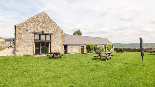 A stone building with a large window and a door, two picnic tables in a grassy area, and a stone wall in the background. - Lathkill Barn (Bakewell)