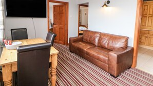 A living room with a brown leather sofa, a wooden dining table with chairs, a TV mounted on the wall, and a wooden door leading to another room. - Haddon Cottage (Bakewell)