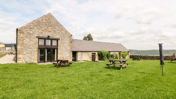 A stone building with a sloped roof, a large window, and a grassy yard with picnic tables. - Ashford Cottage (Bakewell)