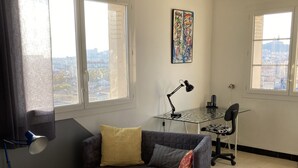 Ein Wohnzimmer mit einem dunkelgrauen Sofa, einem weißen Couchtisch, einem Holzstuhl, einem Schreibtisch mit Computer und Lampe sowie großen Fenstern mit Blick auf die Stadt.