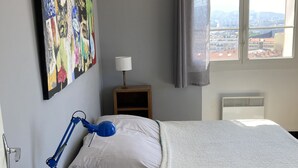Ein Schlafzimmer mit Bett, Schreibtisch mit Lampe, einem Bild an der Wand, einem Fenster mit Aussicht und einer Heizung.