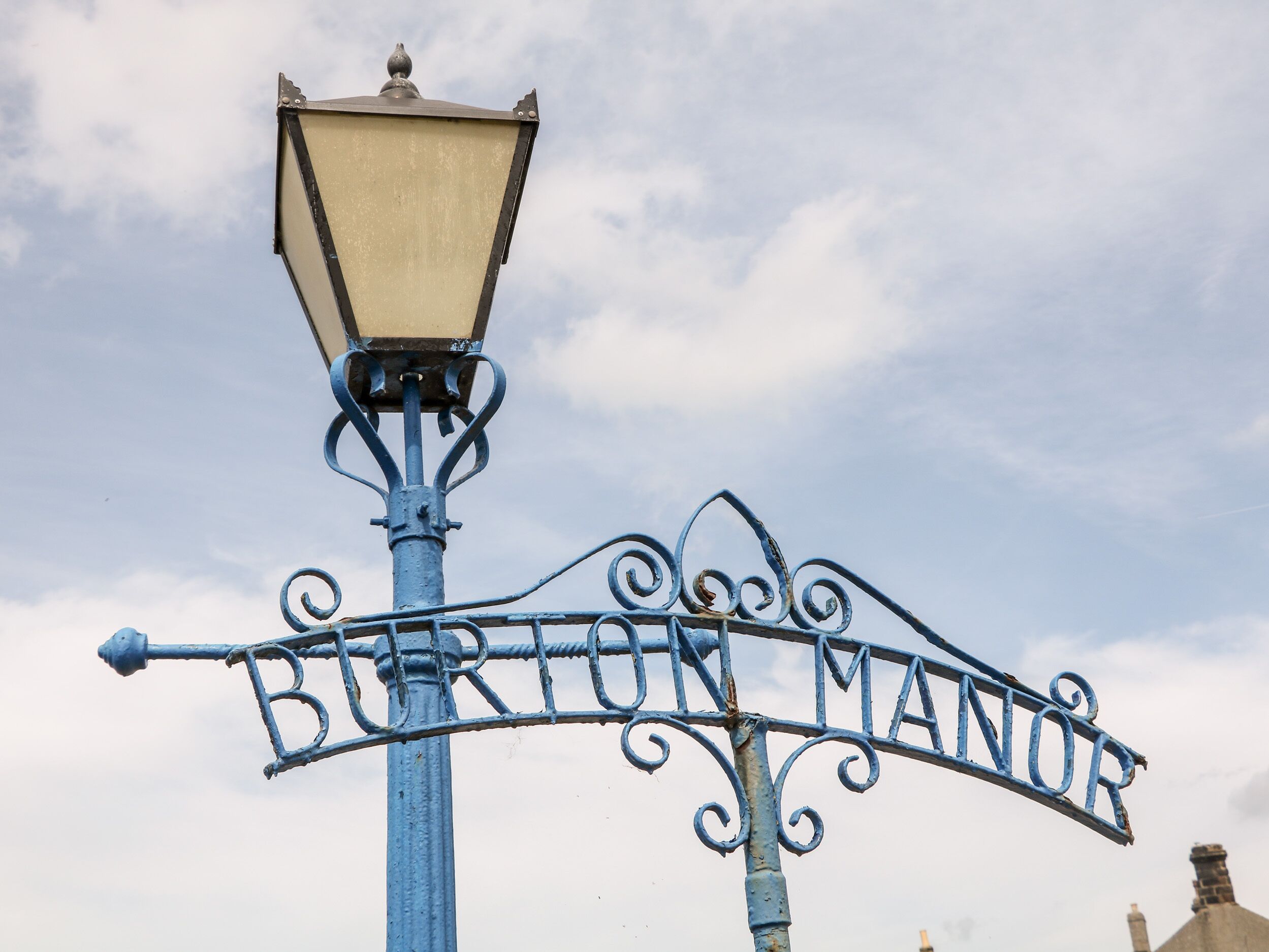 Uma placa de rua com o nome 'BURTON MANDER' fixada num poste de metal azul, com um lampião decorativo acima.