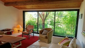 Ein gemütliches Zimmer mit Holzbalkendecke, einem großen Fenster mit Blick auf Grünflächen, einem roten Teppich, einem weißen Sofa, einem Holztisch und einem Stuhl.