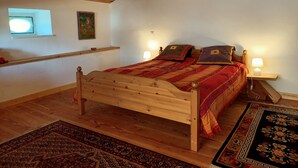 Ein hölzernes Bett mit einer rot-beigen Tagesdecke, zwei Kissen, einem Nachttisch mit Lampe, einem kleinen Fenster und einem Gemälde an der Wand.