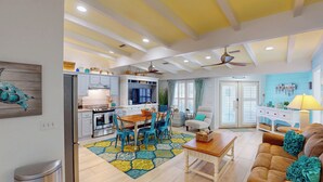 A modern kitchen with white cabinets, a dining area with a wooden table and blue chairs, and a living room with a brown sofa and teal cushions. - Lighthouse Unit B - 2 bedrooms, 2 bathrooms (Jekyll Island)