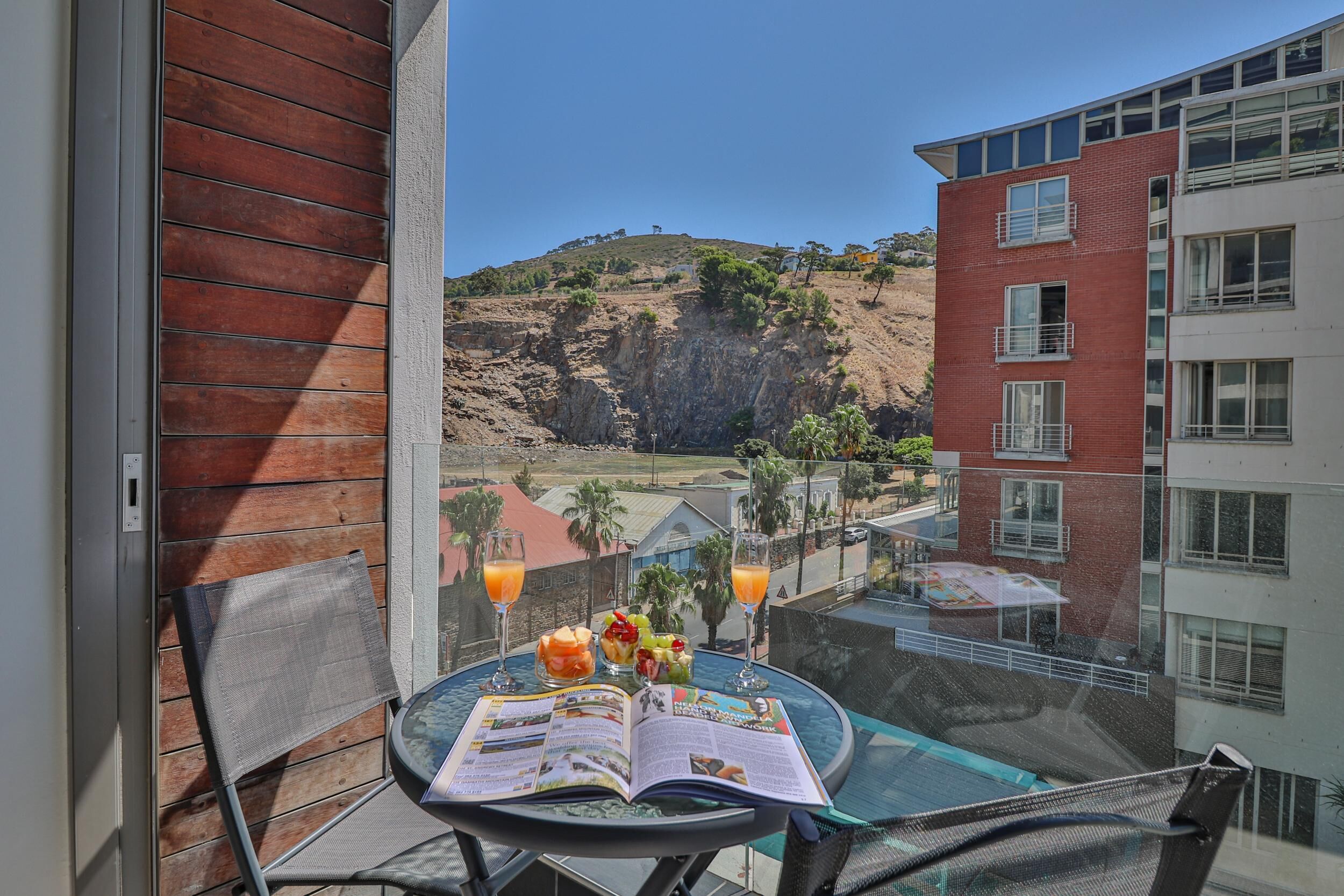 Een balkon met een tafel, waarop een menu, twee sinaasappelsappen en een boek staan. Op de achtergrond is een gebouw met een rode baksteenvoorgevel en een heuvel te zien.