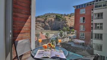 Ein Balkon mit Tisch, auf dem eine Speisekarte, zwei Orangensäfte und ein Buch liegen. Im Hintergrund ist ein Gebäude mit roter Backsteinfassade und ein Hügel zu sehen.