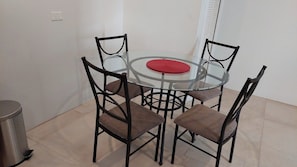 Uma área de refeições com mesa de vidro, quatro cadeiras, um jogo americano vermelho e uma lixeira.