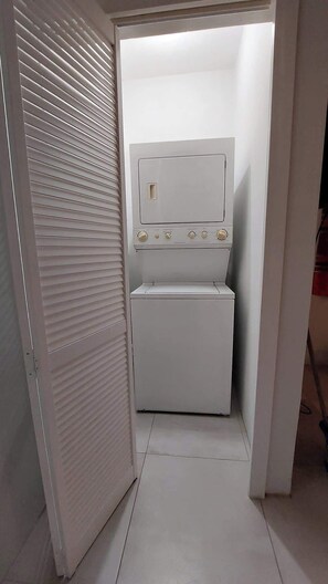 A compact laundry room with a built-in washer and dryer, a small window, and a door with horizontal blinds. - Santa Fe (Port of Spain)