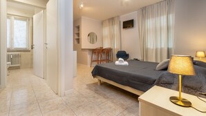 A hotel room with a bed, bedside table, lamp, chairs, a TV, and a bathroom with a radiator.
