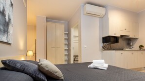 A bedroom with a bed, pillows, a neatly folded towel, a wardrobe, and a kitchenette with a microwave and stove.