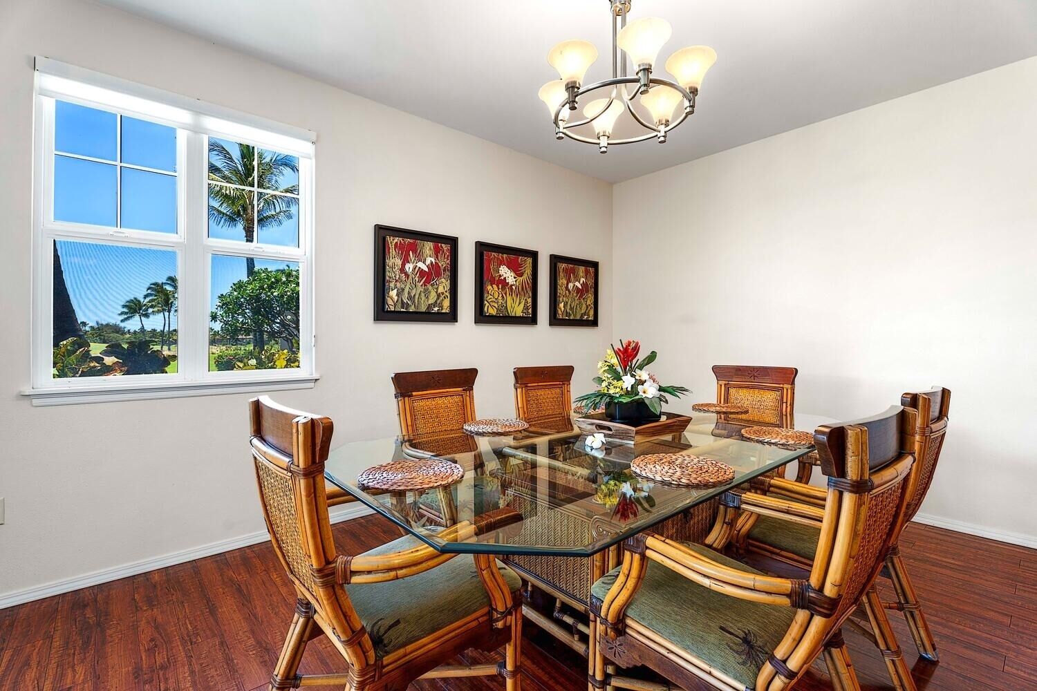 A dining area with a glass table surrounded by bamboo chairs, a chandelier, and framed artwork on the wall.