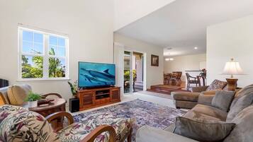 A spacious living room with a large flat-screen TV, comfortable seating, and a view of the outdoors through large windows.