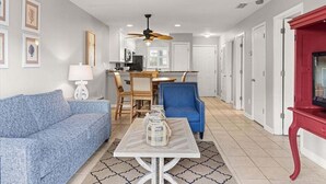 A living room with a blue sofa, a coffee table, and a red entertainment unit. There is a dining area with a table and chairs, and a kitchen in the background. - Romar Beach 112 2 Bedroom Condo by RedAwning (Orange Beach)