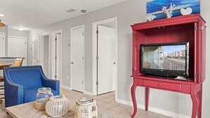 A living room with a red entertainment unit, a blue armchair, and a coffee table with decorative jars. - Romar Beach 112 2 Bedroom Condo by RedAwning (Orange Beach)
