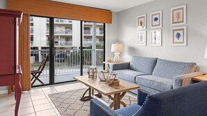 A modern living room with a sofa, a coffee table, and a lamp. There are framed pictures on the wall and a view of a building outside. - Romar Beach 112 2 Bedroom Condo by RedAwning (Orange Beach)