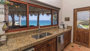 A kitchen with a large window overlooking a beach, granite countertops, wooden cabinets, and a sink. - Costa Azul Condo - Beach front and Ocean Views (San Jose Del Cabo)