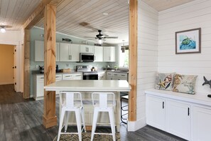 A kitchen with white cabinets, a wooden island with two white bar stools, a ceiling fan, and a framed picture on the wall.