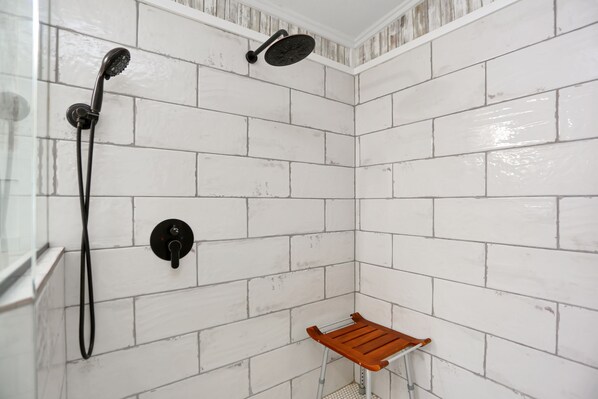 A shower area with white tiled walls, a wooden bench, and two handheld showerheads.