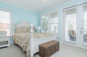 Een slaapkamer met een wit bed, een rieten bankje en een deur met zonwering.