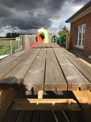 A wooden picnic table with benches, a brick building, and a playground in the background. - Cozy Vacation Home for 8 Persons in Heers (Heers)