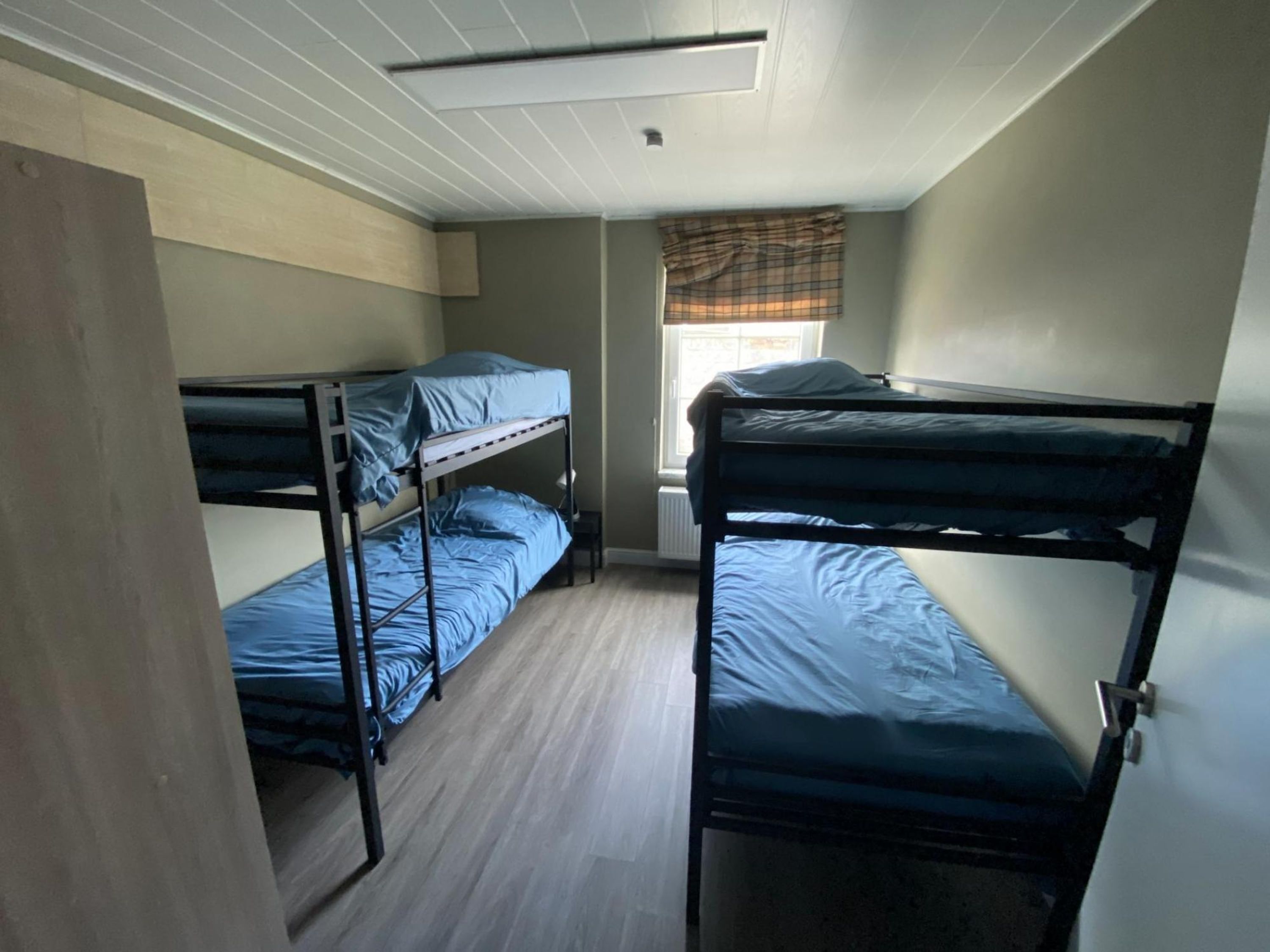 A dormitory room with bunk beds, a window with a plaid curtain, and a radiator under the window.