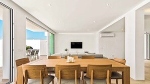 A modern dining area with a wooden table and chairs, a flat-screen TV mounted on the wall, and an air conditioning unit.