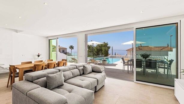 A modern living room with a grey sectional sofa, a wooden dining table with chairs, and a large glass door leading to an outdoor pool area.