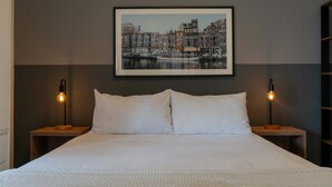 Un lit bien fait, avec du linge de lit blanc, deux tables de chevet avec des lampes et, au mur, un tableau représentant une vue de la ville.