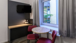 A small room with a wooden table and red chairs, a flat-screen TV mounted on the wall, and a window with curtains.