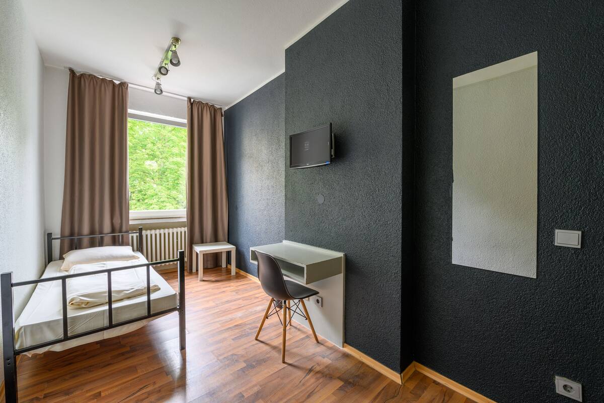 Een kamer met een bed, bureau, stoel, raam met gordijnen, en een aan de muur bevestigde televisie.