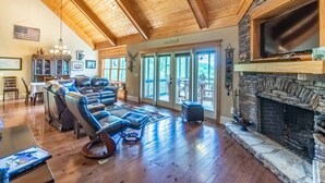 A spacious living room with a stone fireplace, wooden ceiling beams, and a large window overlooking a deck. - Heaven South-Mountain Views - Hot Tub - Fire Pit -Secluded (Bryson City)