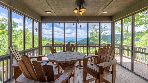A wooden deck with a round table and chairs, large windows offering a view of green hills. - Heaven South-Mountain Views - Hot Tub - Fire Pit -Secluded (Bryson City)