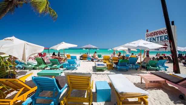 Une plage avec des chaises longues et des parasols aux couleurs vives, un panneau indiquant un centre commercial, et un ciel bleu dégagé.