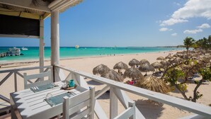 Une vue sur la plage depuis une terrasse en bois, avec une table dressée pour le dîner, donnant sur une plage de sable fin, agrémentée de parasols en chaume et bordée par une mer d’un bleu limpide.