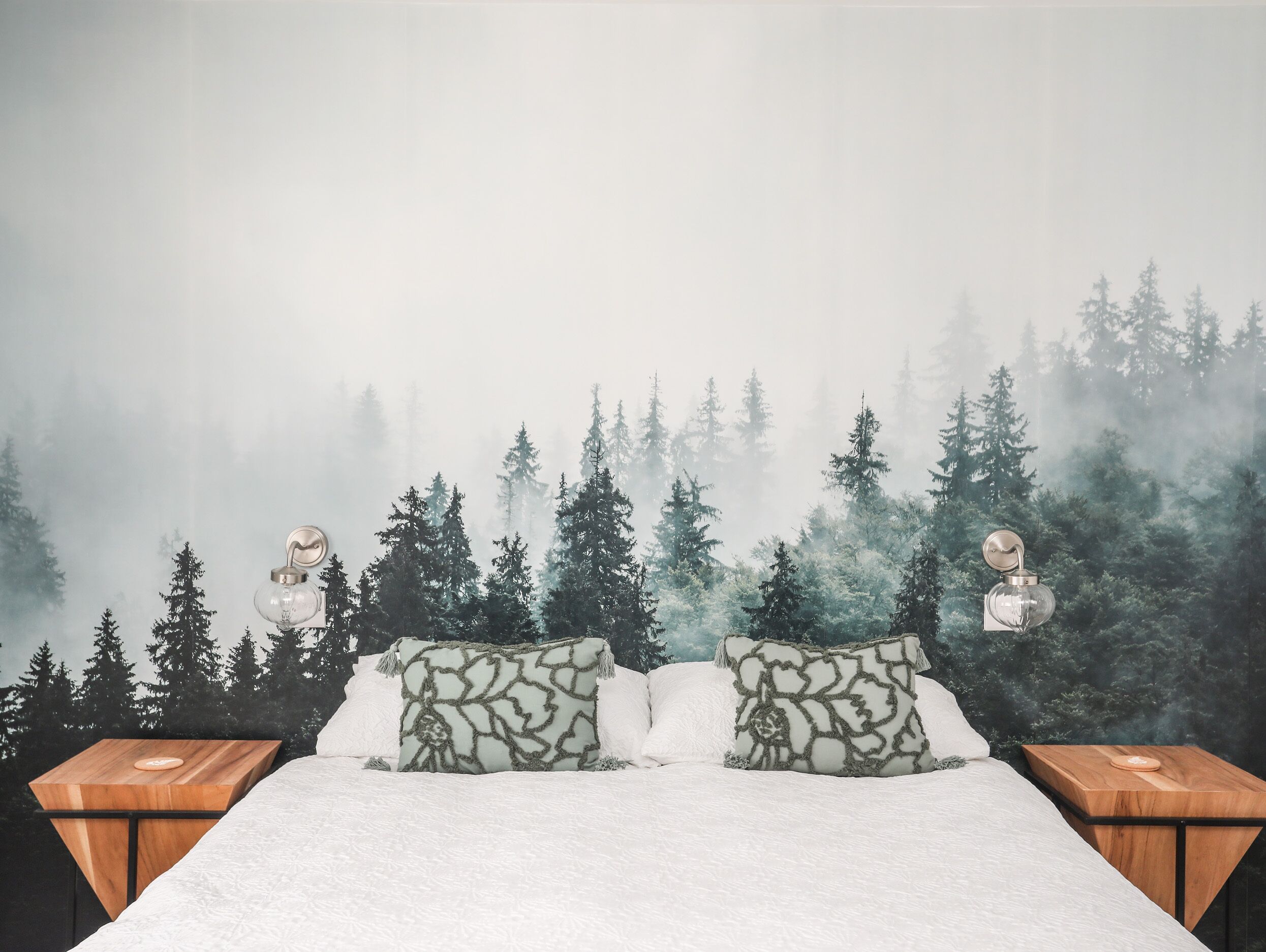 A bedroom with a bed, two nightstands, and a forest-themed wall mural.